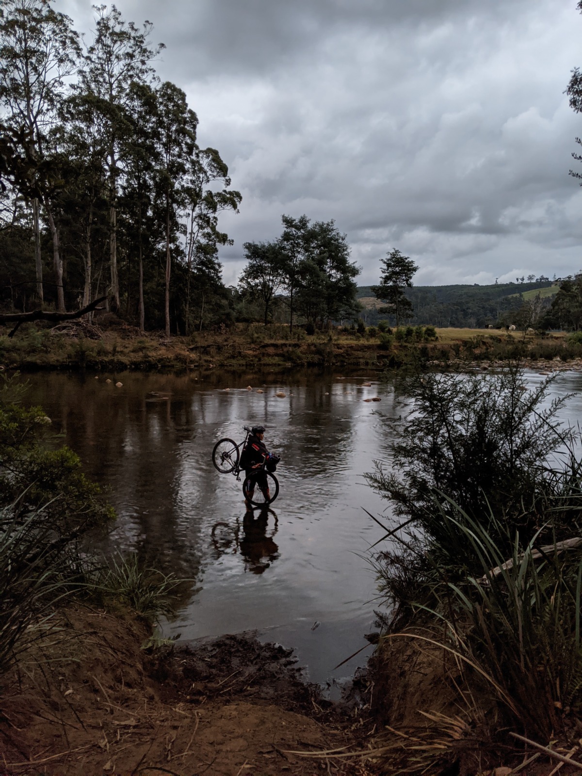 Rider carrying a titanium bike across a river at dusk